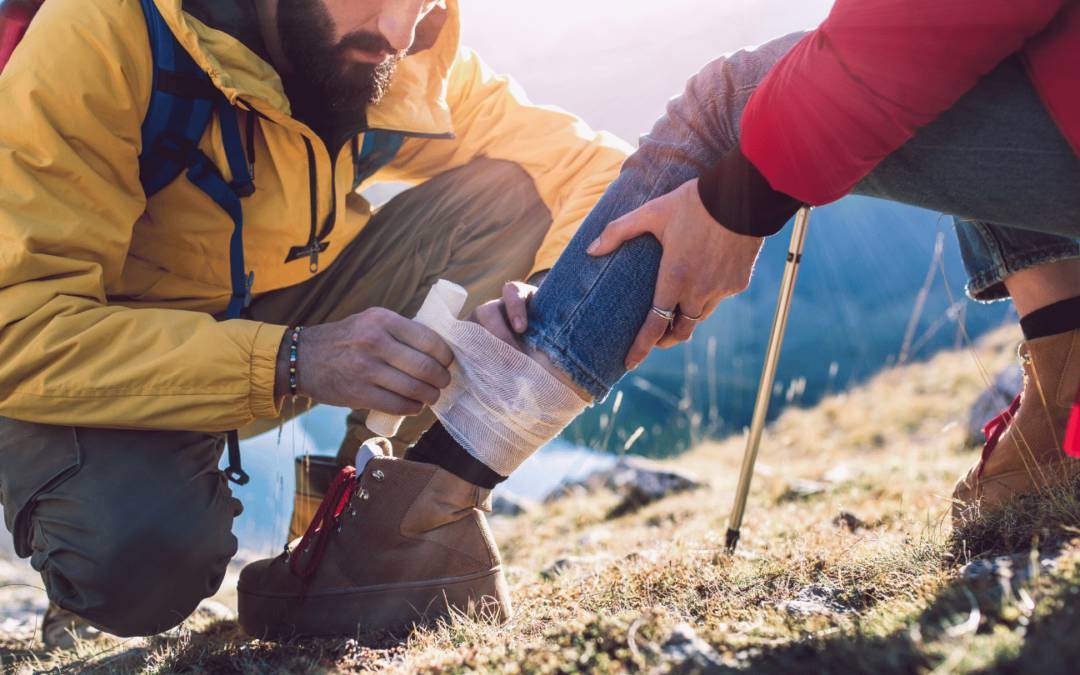 A hiker with first aid training wrapping a leg injury for another hiker on a trail, demonstrating outdoor emergency preparedness.