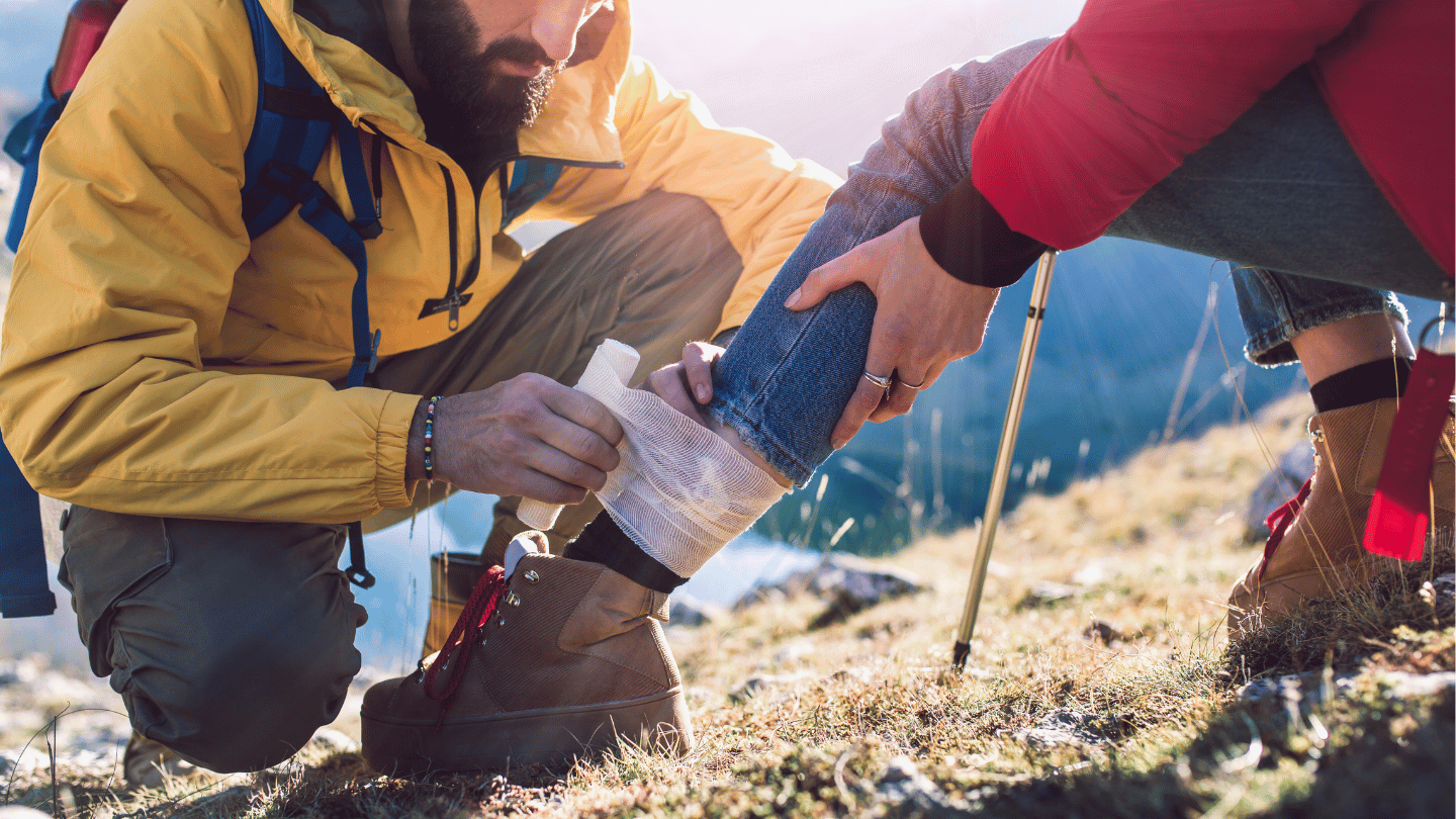 A hiker with first aid training wrapping a leg injury for another hiker on a trail, demonstrating outdoor emergency preparedness.